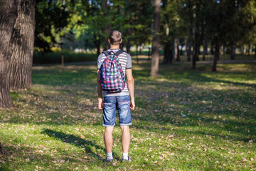 Young guy standing in park with backpack at sunset back view. Traveler, hipster standing in the woods, Hiking, Forest, Journey.