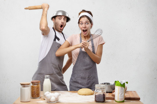 Picture of shocked professional restaurant workers have rivalry or competiton with each other, hold kitchen supplies, fight in funny manner, being surprised or scared to see chef, isolated over white