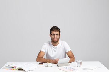 Convinced Caucasian man with stubble and trendy hairstyle, wears casual t shirt, sits at white table, works on creating new project, uses literature and modern device, has serious expression
