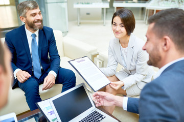 Attractive mixed race assistant manager holding contract for signature in hands while conducting productive negotiations of business partners, interior of spacious boardroom on background