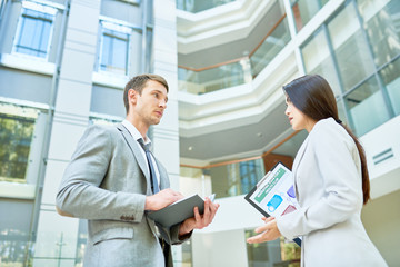 Low angle view of confident young manager presenting her point of view concerning start-up project while having informal meeting with colleague, interior of modern office lobby on background