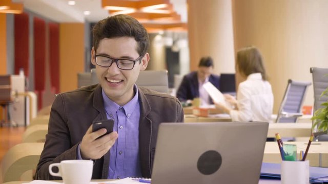 young happy man gets great news about success and triumph during busy working day with papers sitting at table in co working colorful business office space