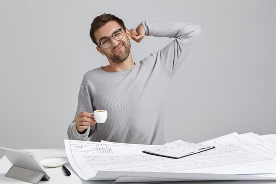 Indoor picture of overworked male creative designer, stretches as sits at table, holds cup of hot drink, has backache after long sitting in workplace, wants to have good rest. Tiredness concept