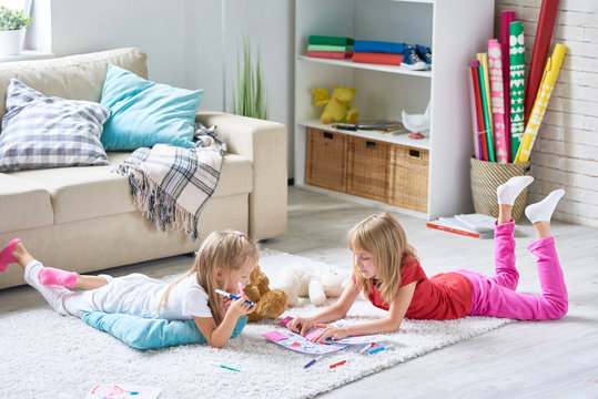 Portrait of cute little sisters coloring pictures together lying on floor on thick plush carpet in cozy living room at home