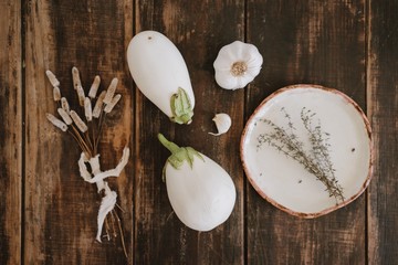 Close up top view of still life with white eggplants with garlic, dried herbs, field flowers and ceramic handmade plate on rustic dark brown wooden table background.
