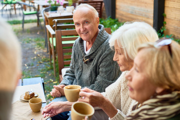 Portrait of handsome senior man with deep blue eyes listening to his friend with interest while having good time together at outdoor cafe, pretty women sitting next to him and enjoying fragrant coffee