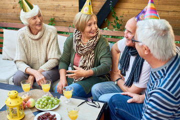 Happy senior people wearing party hats celebrating birthday of their best friend while gathered together at cozy small patio