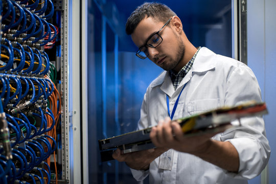 Portrait of young scientist wearing lab coat holding blade server while working with supercomputer in research center