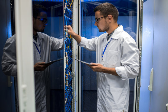 Side view portrait of young man wearing lab coat working with supercomputer connecting blade server cables and  checking data on digital tablet
