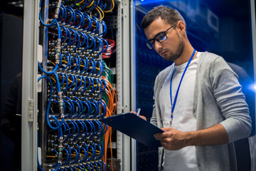 Portrait of young network engineer standing by server cabinet while working with supercomputer in...