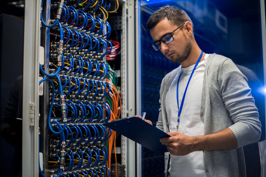Portrait of young network engineer standing by server cabinet while working with supercomputer in data center and holding clipboard
