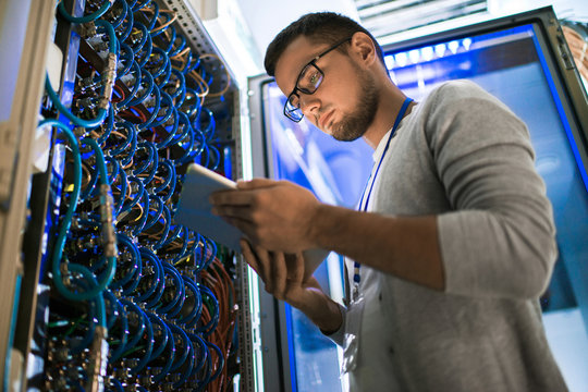 Low angle  portrait of young man using digital tablet standing by server cabinet while working with supercomputer in blue light