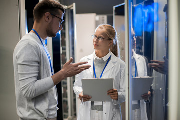 Portrait of  two scientists, man and woman, standing by server cabinets and discussing data while...