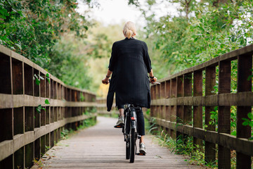 Young stylish blonde woman in black cardigan riding on bicycle on wooden road in park. Handsome girl having a ride. Back view