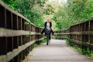 Fototapeta na wymiar Young stylish blonde woman in black cardigan riding on bicycle on wooden road in park. Handsome girl having a ride.