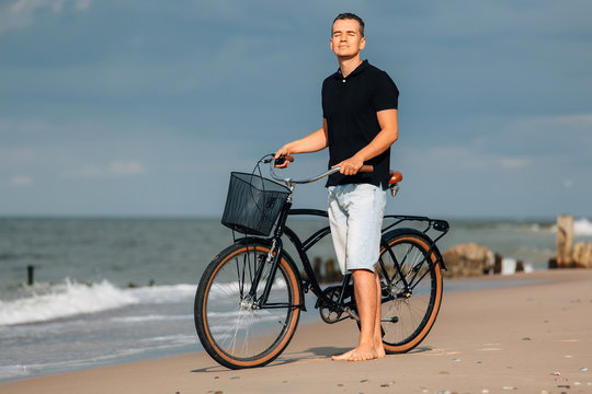 Pretty young man in casual walking on beach with retro bicycle. Handsome guy resting near sea