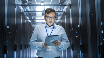 IT Technician Walks Through Rows of Server Racks in Data Center. Simultaneously He Works on a Tablet Computer.