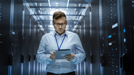 IT Technician Walks Through Rows of Server Racks in Data Center. Simultaneously He Works on a...