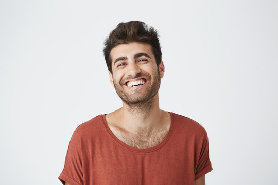 Attractive unshaven young dark-skinned male in red tshirt widely smiling laughing at funny picture on internet. Positive facial expressions and emotions