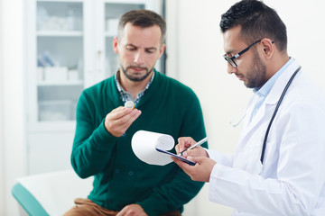 Profile view of confident young doctor in eyeglasses prescribing pills to middle-aged bearded...