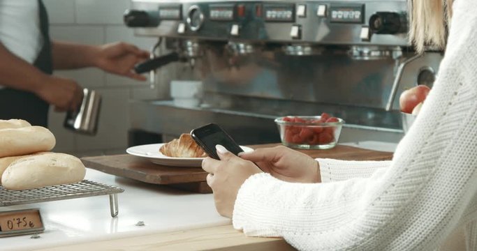 Black barista with tattooed arms serves croissant and coffee to a pretty girl with a smartphone