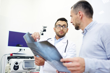 Obraz premium Highly professional radiologist analyzing X-ray images of middle-aged patient and giving necessary recommendations, interior of modern office on background