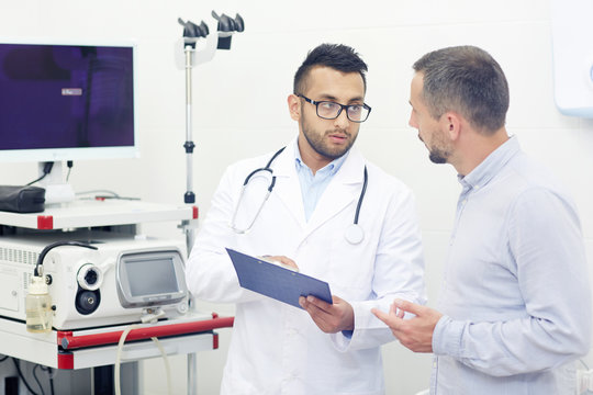 Handsome young doctor in eyeglasses discussing medical test results with male patient and prescribing necessary treatment, interior of modern office on background