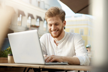 Handsome fashionable european male freelancer with trendy hair and beard sits outside in sunny weather, working on his laptop computer, translating articles.