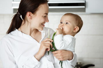 Close up of little cute baby boy eating green onion with funny surprised expression from it taste. Mother smiling and looking at her child.