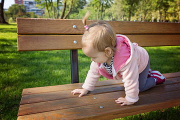 Eighteen months old toddler girl climbing on the wooden bench in the park on a sunny autumn day