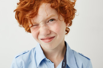 Close up of funny ginger boy with freckles and red cheeks screw up eye because of bright lightning in school class.
