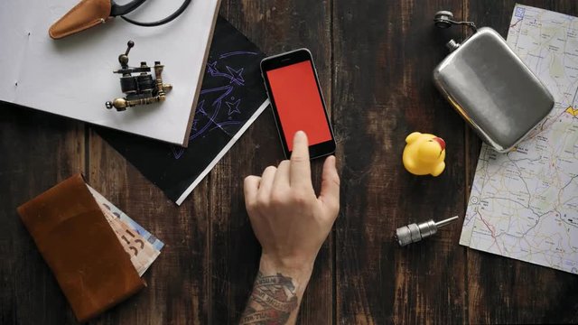 Cool hipster man with artisan tattoos swipes different screens with finger, top view of wooden table, with artifacts and personal items, such as wallet, map, travel essentials, chroma key filled