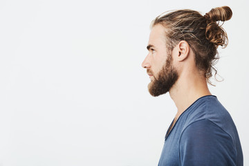 Close up of good-looking bearded hipster guy with hair in bun, in blue t-shirt standing in profile,...