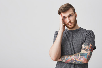 Close up of beautiful unhappy bearded hipster man with tattoo and good hairstyle holding head with...