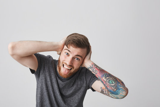 We're All Mad Here. Portrait Of Good-looking Funny European Man With Beard And Tattooed Arm Looking In Camera With Crazy Expression, Clothing Ears With Hands, Showing Tongue And Having Fun.