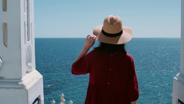 Beautiful young woman in romantic red dress and straw hat stands on top of pier or balcony overlooking blue waters of ocean or sea, wind and summer breeze blows her hair, concept getaway, vacation