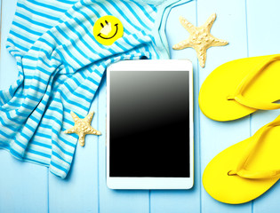Summer women's accessories: sunglasses, yellow step-ins, dress, tablet, camera, on blue wood background.