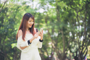 Cute asian woman is reading pleasant text message on mobile phone while sitting in park in warm spring day,gorgeous female listening to music in headphones and searching information on cell telephone.