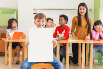 Cute boy holding blank white poster with happy face in kindergarten classroom, kindergarten...