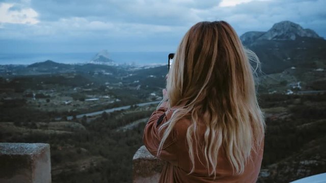 Pretty beautiful blonde selfish woman self centered makes selfie on smartphone while sits on top of car during twilight hours in sunset, beautiful and calm views of independent teenager