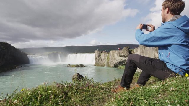 Iceland tourist taking photo with smart phone of waterfall Godafoss. Man taking picture with smart phone camera on travel visiting tourist attractions and landmarks in Icelandic nature on Ring Road.