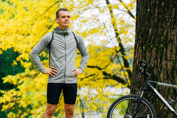 Young pretty athletic man standing with bicycle in autumn park. Fall season background. Male...
