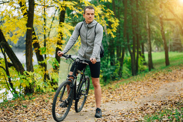 Fototapeta na wymiar Young pretty athletic man standing with bicycle in colorful autumn park. Fall season background. Male cyclist on the road with fallen leaves