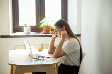 Female employee taking a minute break resting her aching eyes after computer work, having headache . Young tired woman sitting next to office window and pot plants feeling depressed