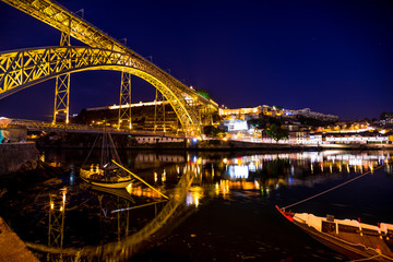 Iconic symbol of Oporto city. Scenic illuminated Dom Luis I bridge with boats reflecting on Douro River at night in Porto, Portugal's second largest city. Picturesque urban skyline.