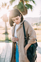 Obraz premium Trendy millennial fashion student or model poses to camera on seaside promenade, looks down in humble and shy way, hand in pocket of trendy cotton coat