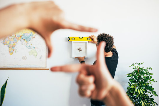 Young couple hangs frame or painting in new home on white wall next to world map, woman puts her fingers in shape of square to frame how the photo will look