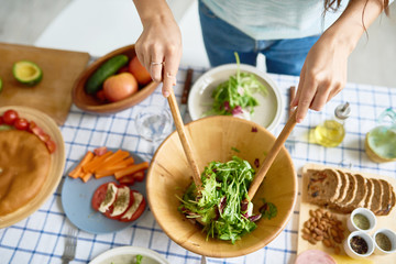 Closeup of female hands mixing green salad in wooden bowl at table with wholesome food while preparing family dinner