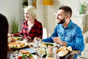 Portrait of happy young man and woman sitting at table with delicious meals, enjoying festive dinner with friends at home gathering