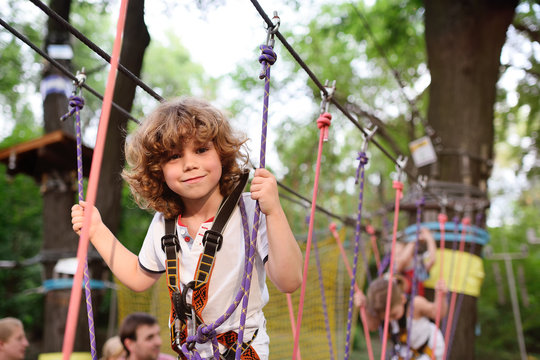 Child - A Cute, Curly-haired Boy In An Amusement Park Climbing The Rope Road. Hyperactive Child